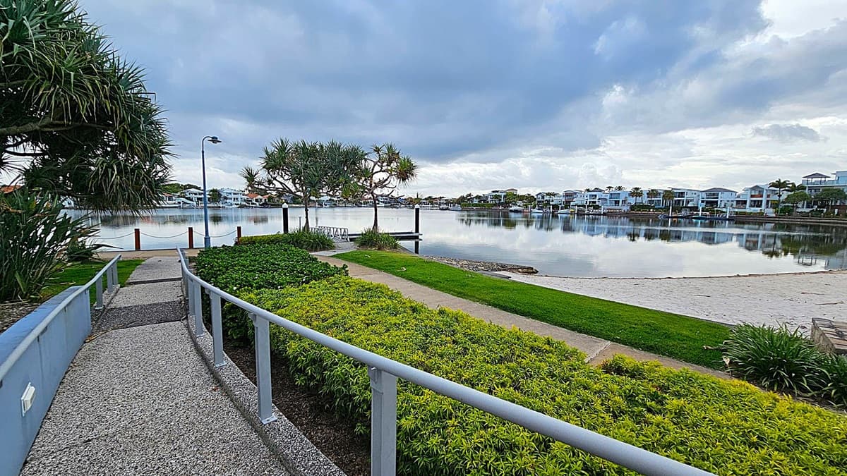A paved path leads through a landscaped area with neatly trimmed bushes and palm trees. On the left, there is a modern building with a flat roof and a small set of stairs. On the right, a ramp with a metal railing ascends towards another building partially obscured by trees. The foreground features a mix of grass and mulch, while the background includes more greenery and architectural elements.