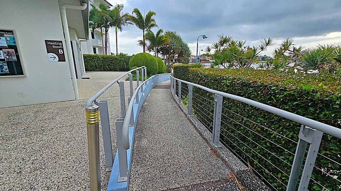 A pathway with a grey, speckled surface curves gently to the right, bordered by a metal railing. On the left, a building with a sign for "Meeting Room B" and a notice board is visible. To the right, a neatly trimmed hedge runs alongside the path, with palm trees and other greenery in the background. A streetlamp and a glimpse of water and buildings can be seen in the distance.