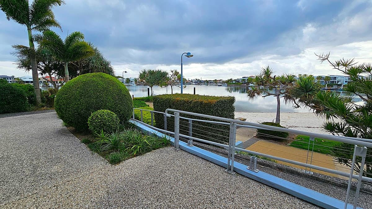 A waterfront scene with a pebble pathway in the foreground, bordered by neatly trimmed bushes and a round, manicured shrub on the left. Tall palm trees rise behind the shrubbery. A metal railing runs along the path, overlooking a calm body of water. Across the water, modern houses are visible. The sky is overcast, casting a grey tone over the landscape.