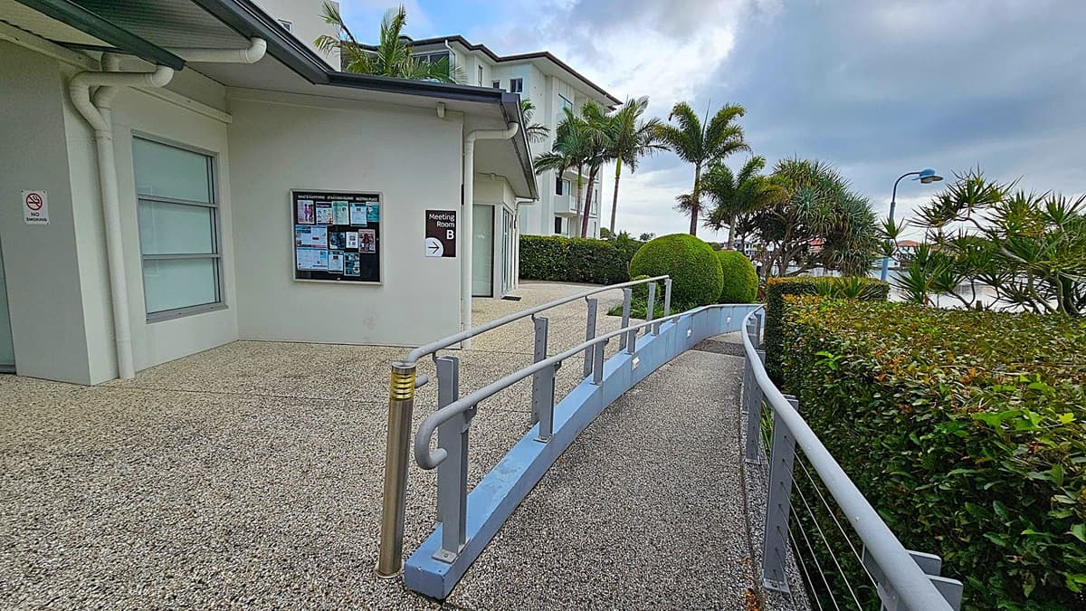 A building with a grey exterior and a window on the left. A notice board and a sign for "Meeting Room B" are mounted on the wall. A ramp with metal railings curves from the foreground to the right, bordered by a hedge. Palm trees and manicured bushes are visible in the background, along with a streetlamp. The ground is covered with a speckled surface.