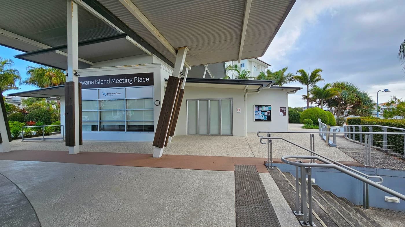 A modern building with a sign reading "Kawana Island Meeting Place" on the left. The building has large windows and a corrugated metal roof. In the centre, there are closed sliding doors and a notice board on the wall. On the right, a pathway with metal railings leads to a staircase. The area is surrounded by well-maintained greenery, including palm trees and bushes. The ground is paved with textured concrete.
