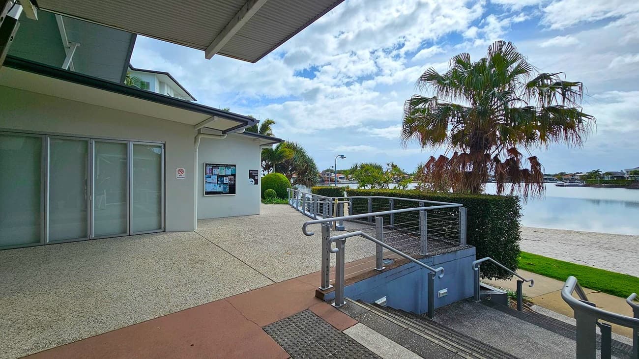 A building with large glass doors on the left, featuring a notice board on the wall. A pathway leads to a railing and steps descending to a lower level. On the right, a palm tree stands near a body of water with a sandy shore. The background includes more trees and distant buildings across the water. The floor is a mix of concrete and textured surfaces, and the ceiling is metal with visible beams.