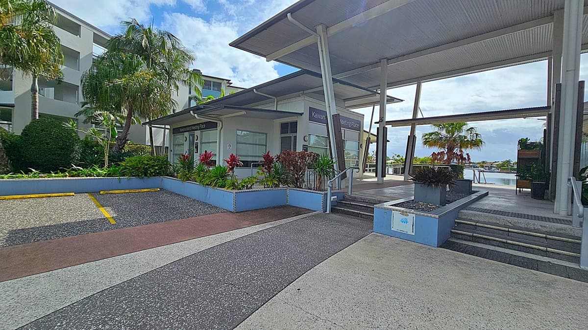 A modern building with a slanted roof and large windows is on the right, labelled "Kawana Island Meeting Place." Steps lead up to the entrance, flanked by planters with red and green foliage. On the left, there are palm trees and a multi-storey building with balconies. The foreground has a textured pavement with yellow parking lines. In the background, there's a glimpse of water and more greenery.
