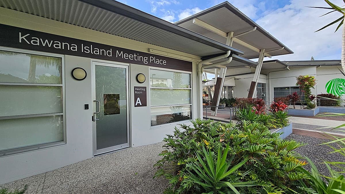 A modern building with a sign reading "Kawana Island Meeting Place" above a door labelled "Meeting Room A". The exterior features large windows and a grey, corrugated roof. In the foreground, there are lush green plants and a textured pathway. To the right, more plants are in raised beds, and a green abstract design is visible on a distant wall. The structure has a contemporary design with angled supports and overhangs.