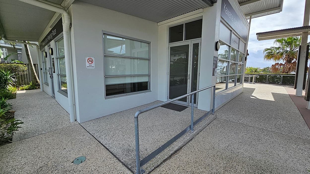 A building corner with large windows and a glass door. A "No Smoking" sign is on the wall. A metal handrail runs along the edge of the concrete path. To the left, there are plants and a fence. On the right, a pathway leads to a railing with palm trees and greenery in the background. The ceiling is corrugated metal.