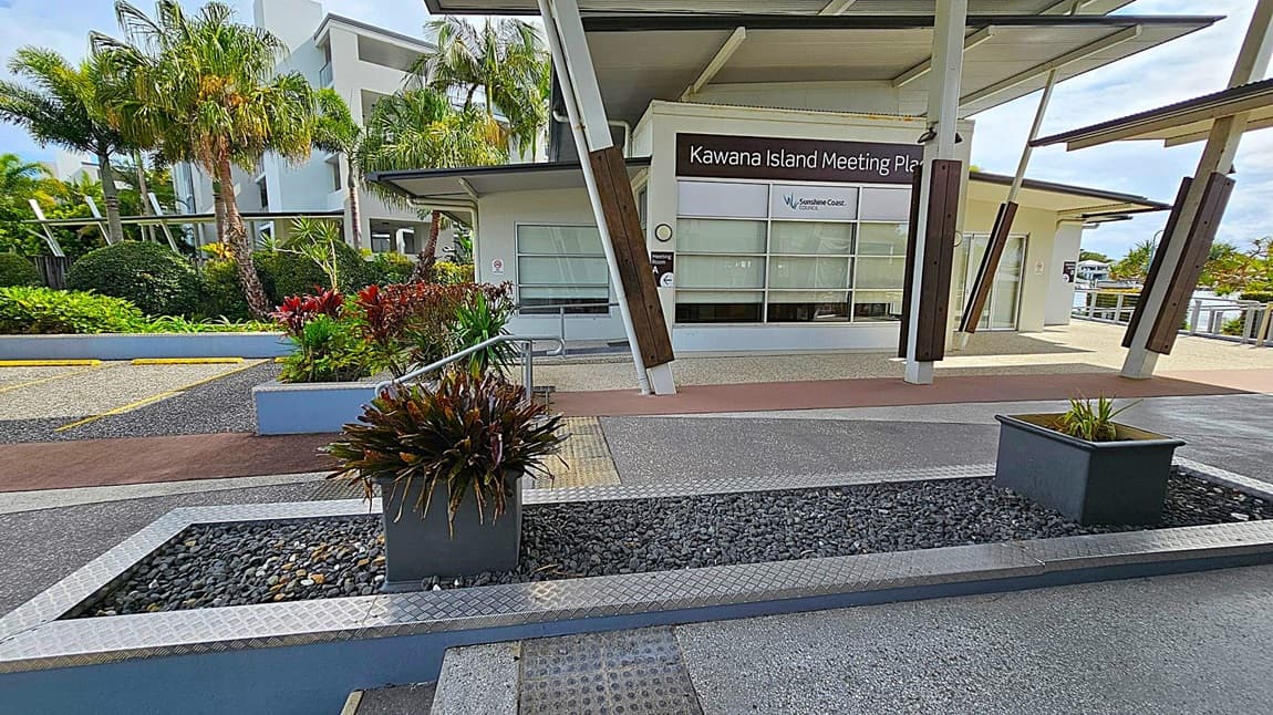 In the foreground, there are two rectangular planters with green plants, set on a bed of dark pebbles. The background features the Kawana Island Meeting Place building with large windows and a sign for the Sunshine Coast Council. To the left, there are lush green plants and palm trees. The ground is a mix of textured concrete and pebble surfaces, and the building has a modern design with angled supports and a flat roof.