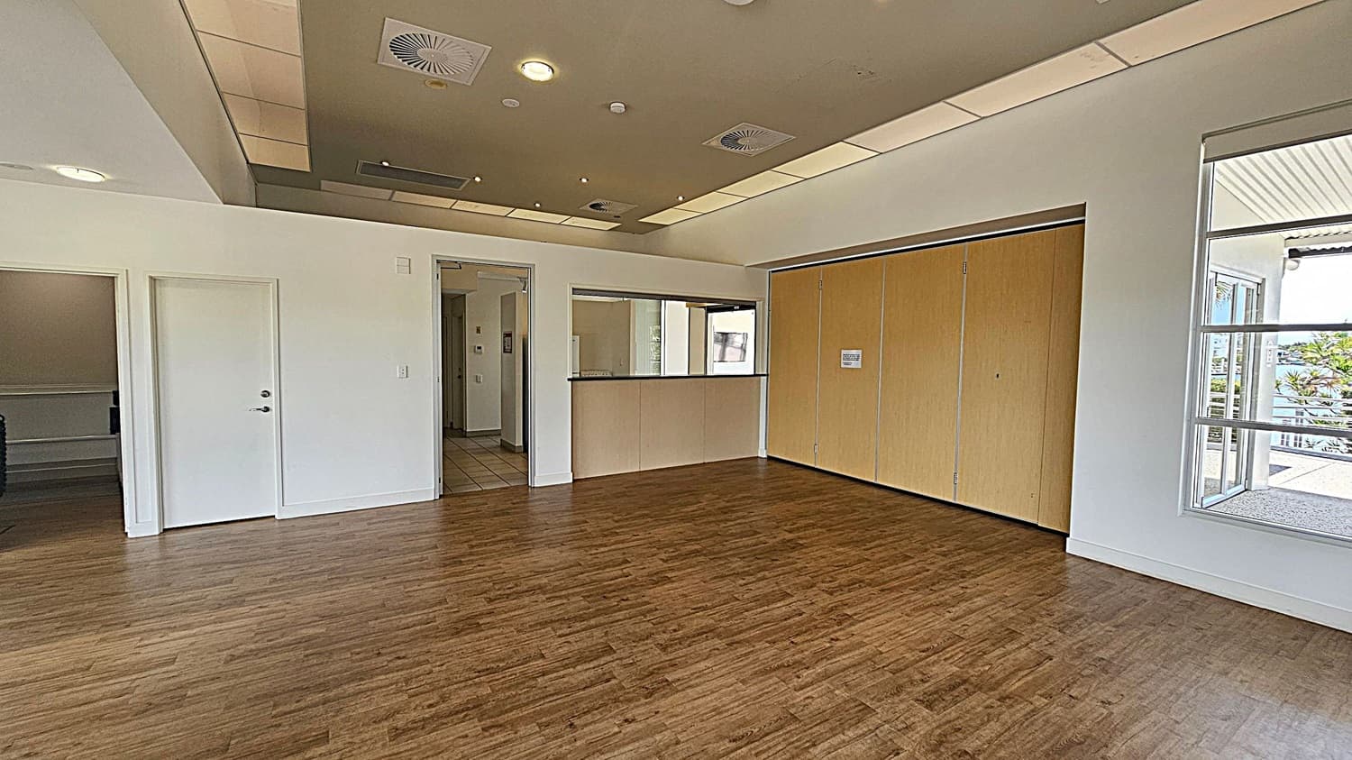 A room with wooden flooring and a light-coloured ceiling featuring recessed lighting and air vents. On the left, there are two white doors, one slightly ajar, leading to another area. In the centre, a counter with a window above it is set into the wall. On the right, large wooden sliding doors are closed, and a window reveals an outdoor view with greenery and a building.