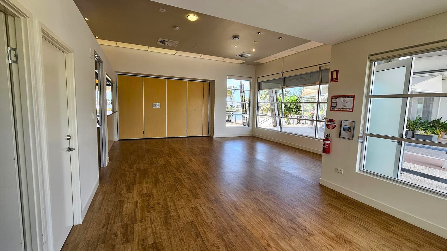 A spacious room with wooden flooring and a light-coloured ceiling featuring recessed lighting and air vents. On the left, there are two closed doors. The back wall has large windows and a set of wooden folding doors. On the right, more windows allow natural light in, with a fire extinguisher and safety instructions mounted on the wall. Outside, there are visible plants and a walkway.