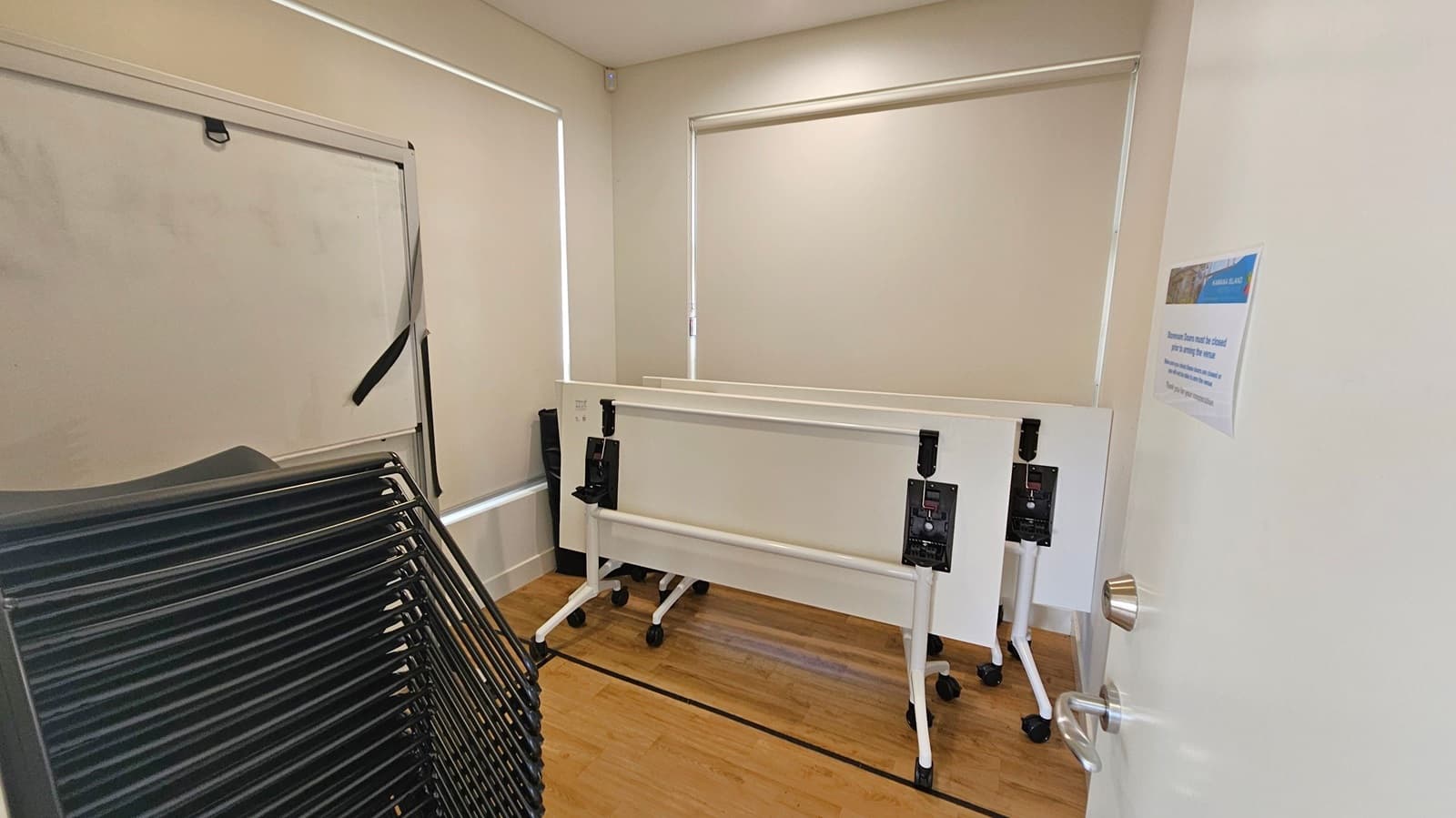 A small room with light-coloured walls and wooden flooring. On the left, there is a stack of black chairs. In the centre, two white folding tables with wheels are positioned against the wall. The windows have closed blinds. On the right, a white door is partially open, with a silver handle and a notice attached to it.