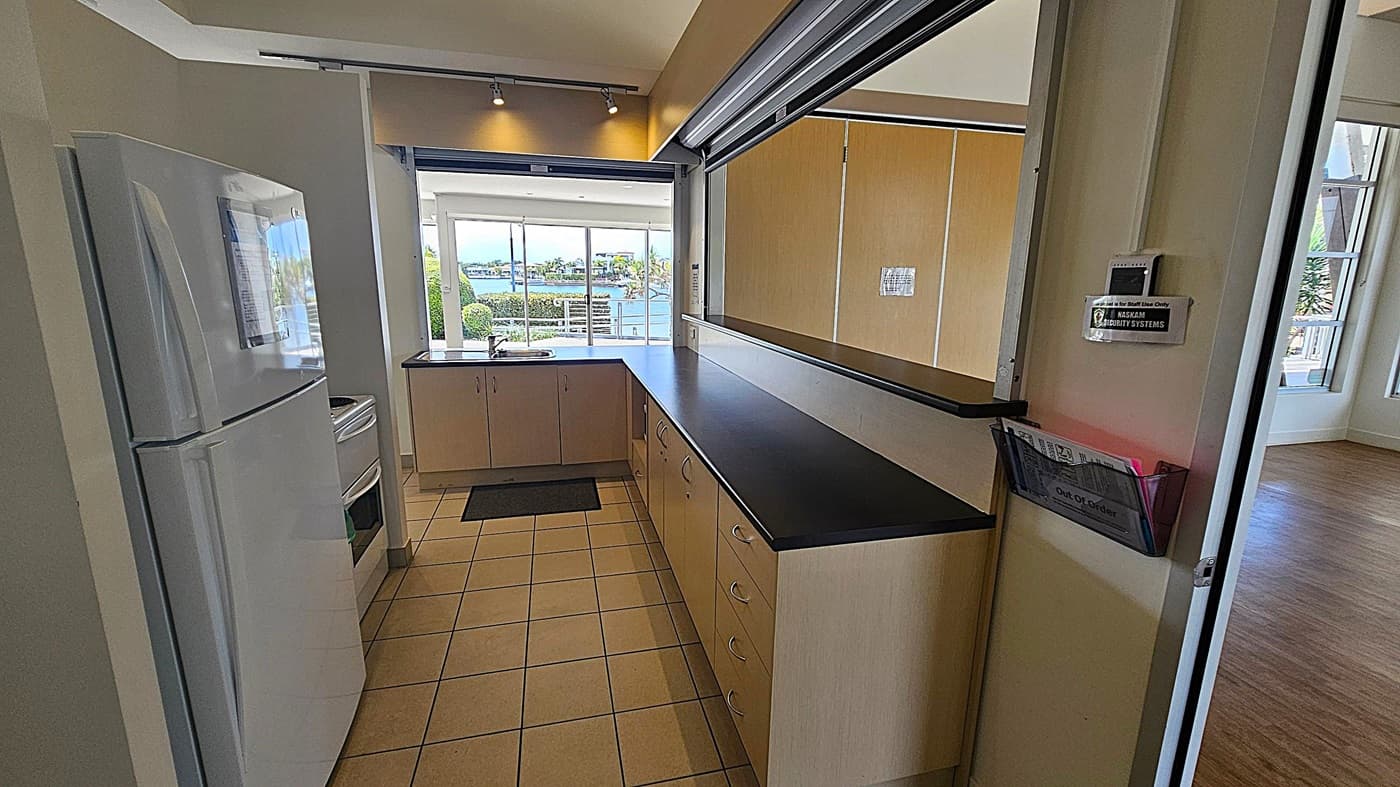 A kitchen with a tiled floor and a black countertop. On the left, there's a white refrigerator and a stove. The countertop extends along the right side with multiple drawers. In the background, large windows offer a view of a waterfront with trees and buildings. A small mat is placed near the sink. A wall-mounted security system and a holder with papers marked "Out of Order" are visible on the right. The ceiling has recessed lighting.