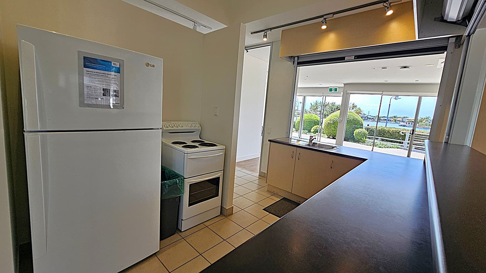 A kitchen area with a white fridge on the left, featuring a notice on its door. Next to it is a white electric stove. The floor is tiled. On the right, a counter with a sink overlooks a large window. Beyond the window, there are neatly trimmed bushes and a view of water and distant buildings. The ceiling has track lighting.