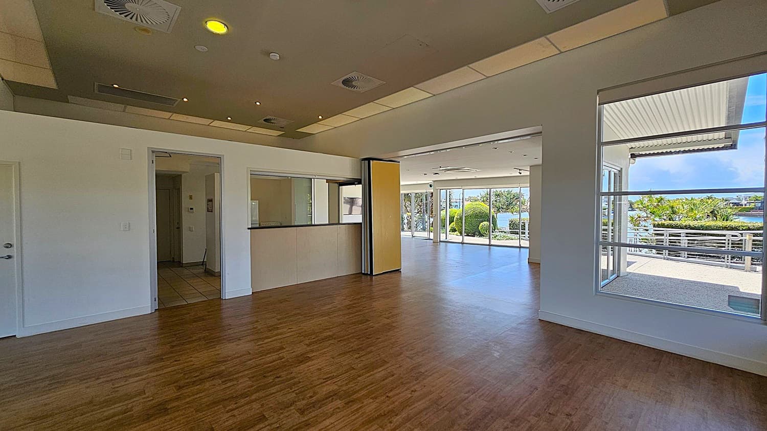 A spacious room with wooden flooring and a ceiling featuring recessed lighting and air vents. On the left, there is a small counter with a window and a doorway leading to another room. The right side has large windows and glass doors opening to an outdoor area with greenery and a view of water. The outdoor area has a railing and a covered section.