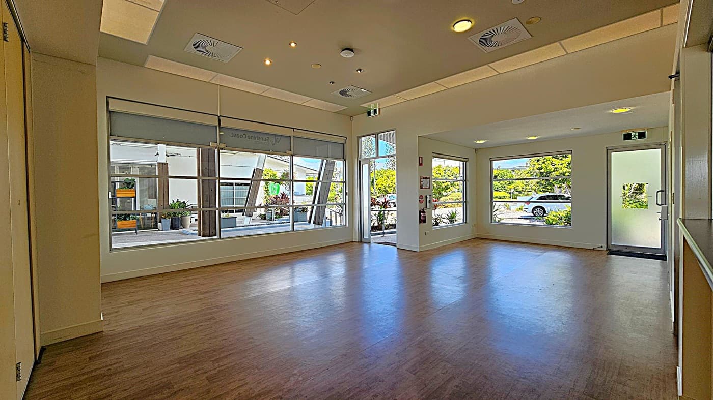 A spacious room with large windows on the left and right, allowing natural light to fill the space. The windows reveal a view of greenery and a parked car outside. The floor is wooden, and the ceiling features recessed lighting and air vents. A glass door on the right leads outside, and there are potted plants visible through the windows. The room has a clean, modern appearance.