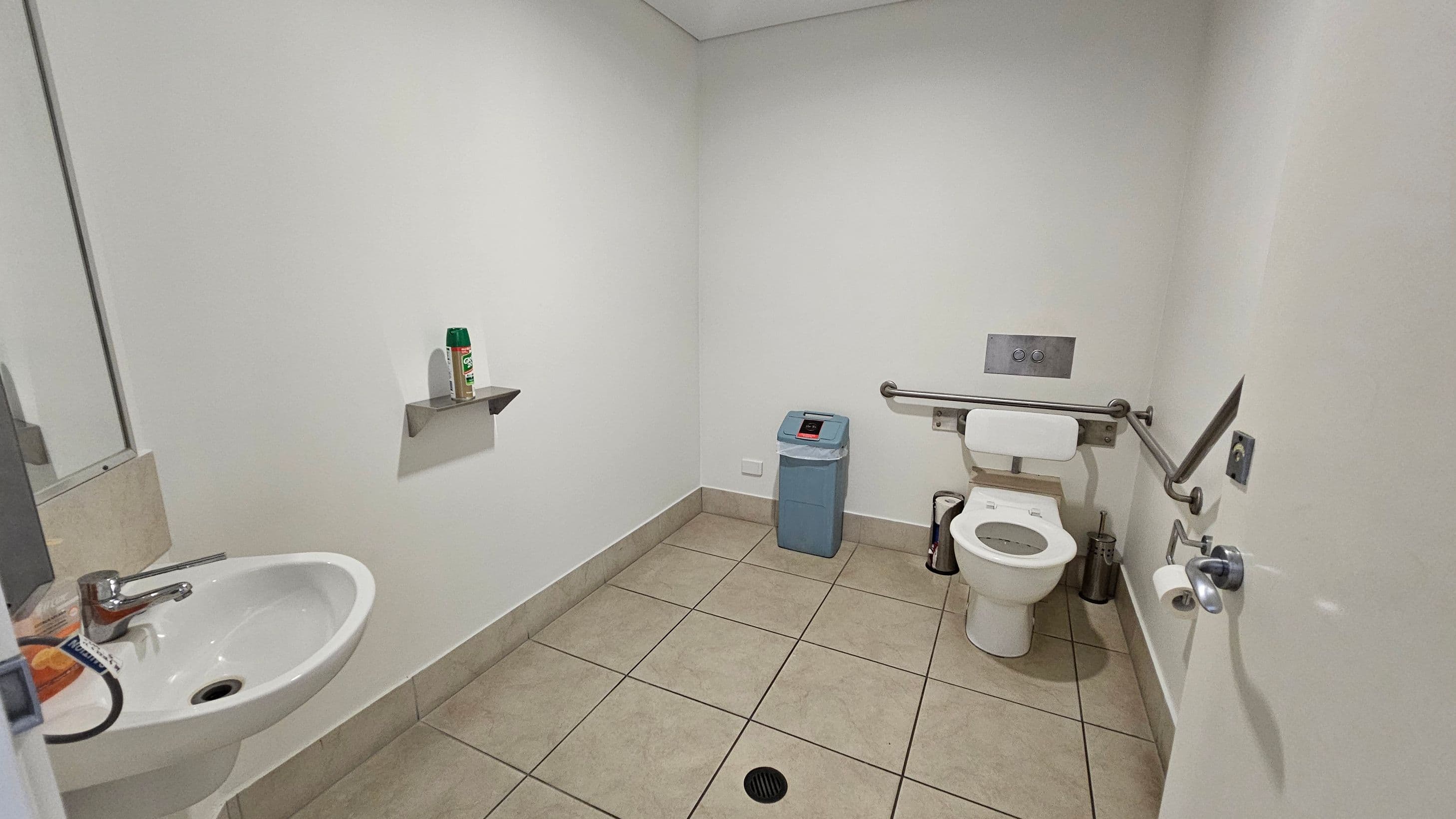 A bathroom with beige tiled flooring and white walls. On the left, there's a white sink with a silver tap and a mirror above it. A small shelf holds a can of spray. In the centre, a blue bin is placed against the wall. On the right, a toilet with a backrest is equipped with a grab rail. A toilet roll holder is mounted on the wall, and a small bin is nearby. The ceiling is plain white.