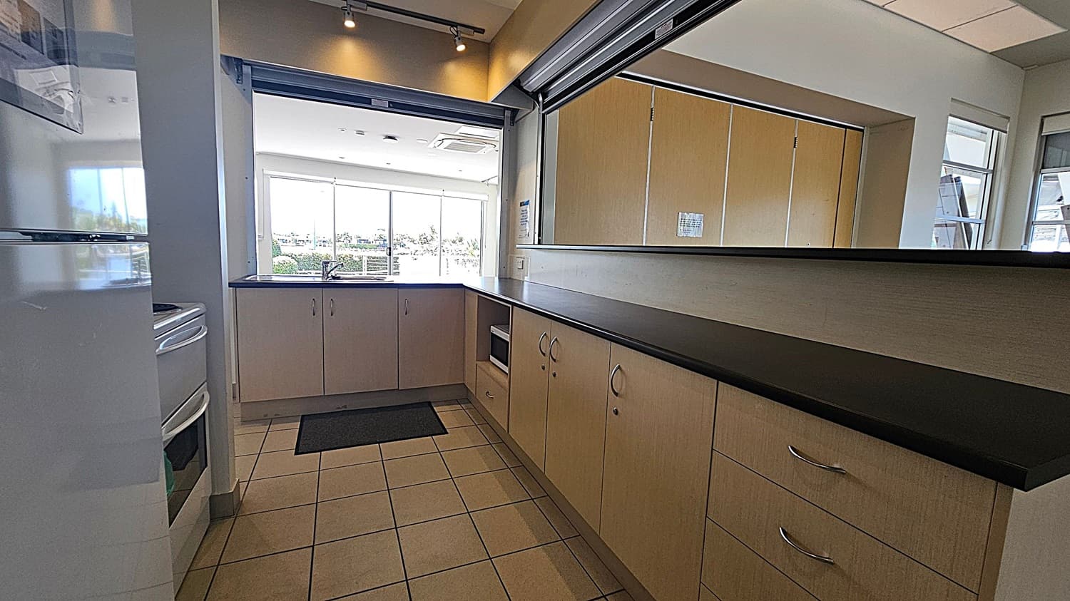 A kitchen area with a stainless steel sink and drainer on the left, set into a dark countertop. Below the counter are light-coloured cabinets and a microwave. On the right, a white wall features an Automated External Defibrillator (AED) in a wall-mounted case. In the background, large windows and glass doors reveal an outdoor area with plants and a pathway. The floor is tiled, and the ceiling is not visible.