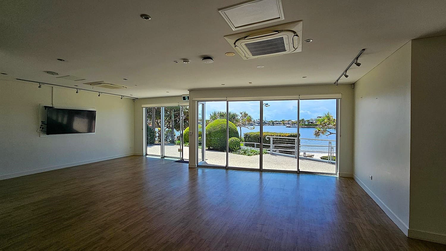 A spacious room with wooden flooring and a white ceiling featuring recessed lighting and an air conditioning unit. On the left wall, a flat-screen TV is mounted. The right side has large glass sliding doors leading to an outdoor area with neatly trimmed bushes and a view of a body of water and distant buildings. The room is well-lit by natural light coming through the glass doors.