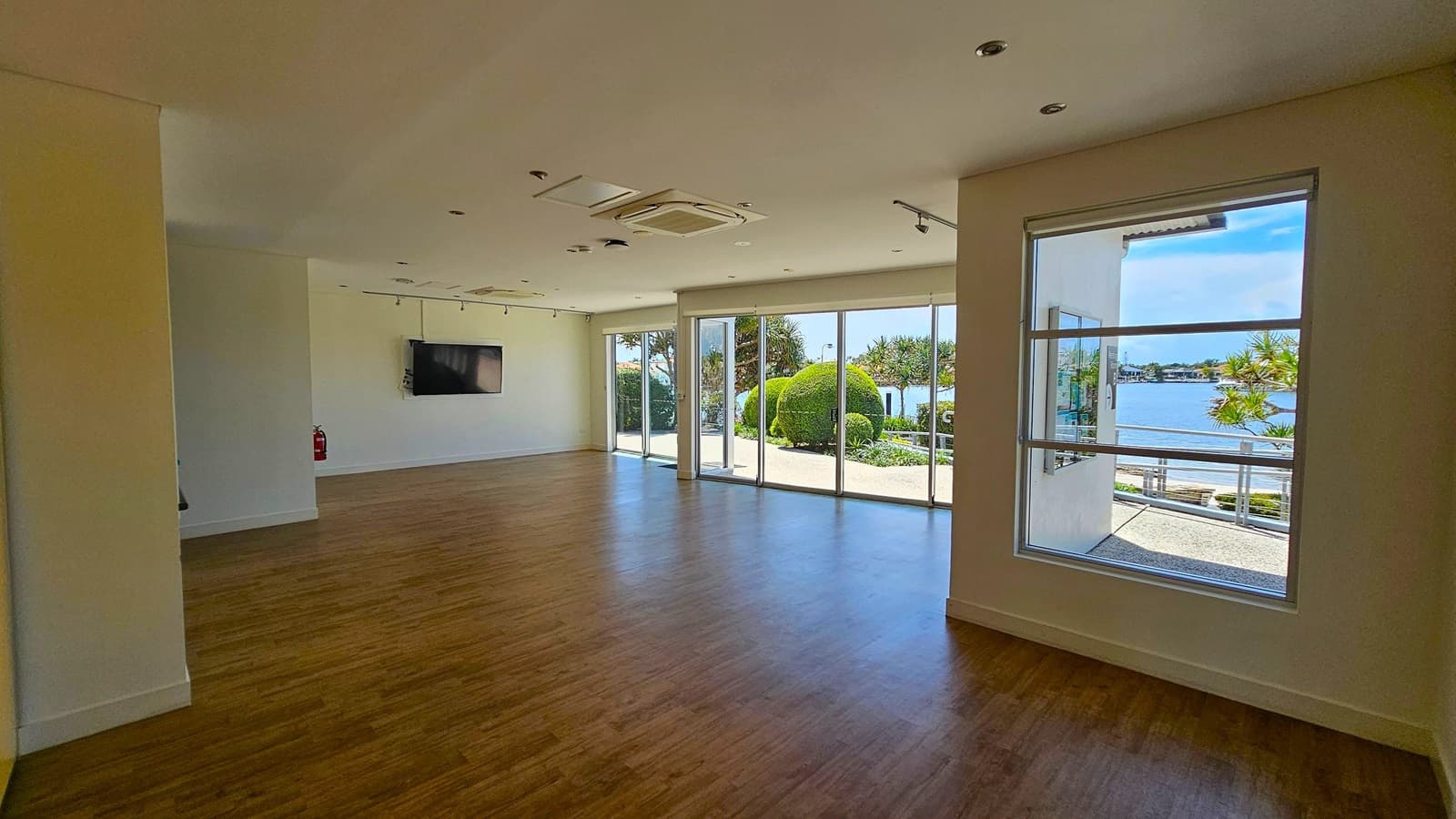 A spacious room with wooden flooring and a white ceiling featuring recessed lights and an air conditioning unit. On the left, a wall-mounted television and a fire extinguisher are visible. The right side has large windows and glass doors, offering a view of a garden with neatly trimmed bushes and a body of water in the background. The room is well-lit with natural light streaming in through the windows.