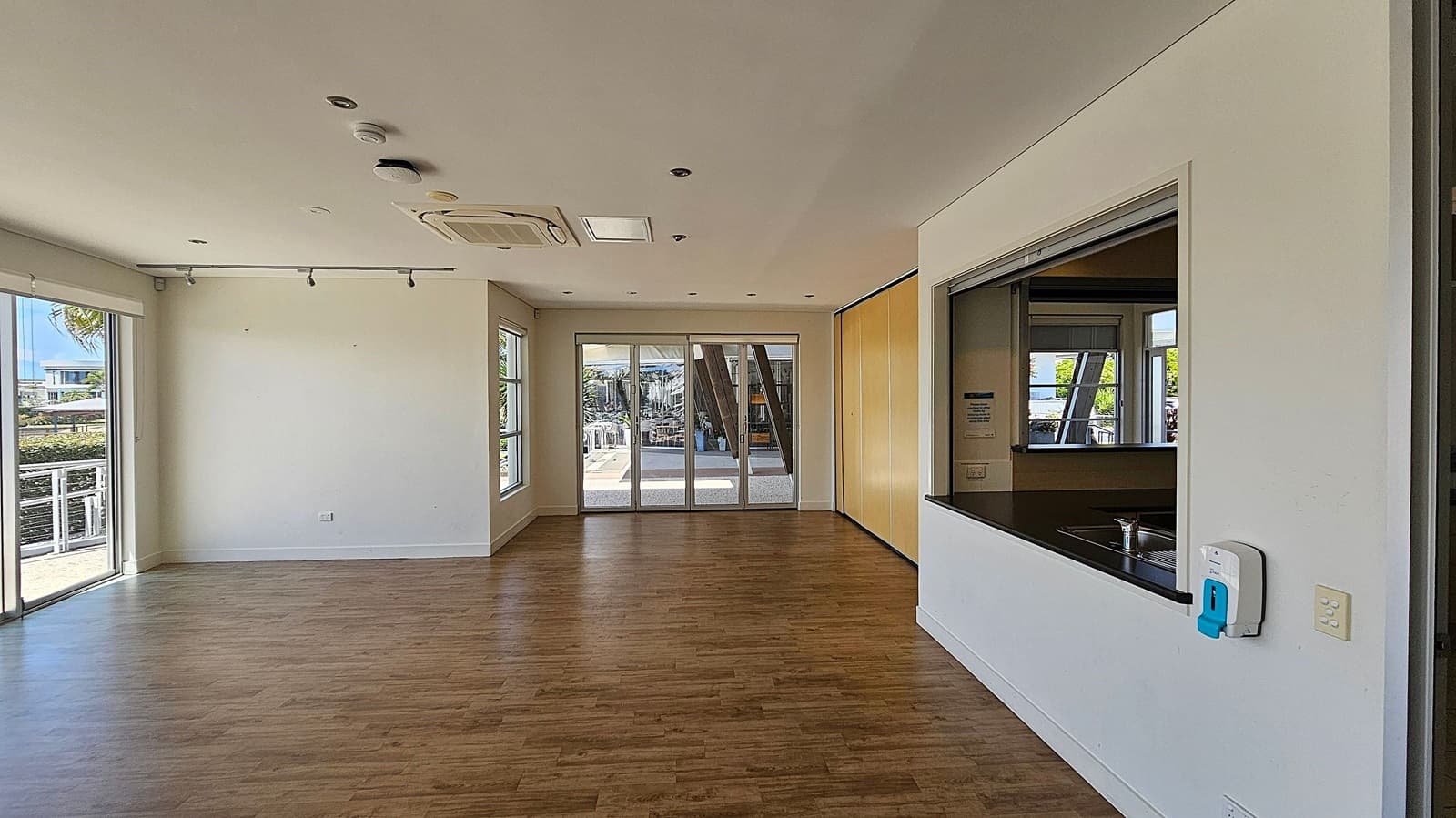 A spacious room with wooden flooring and white walls. On the left, large windows and a sliding glass door provide natural light and a view outside. In the centre, a set of glass doors leads to an outdoor area. On the right, there's a serving hatch with a countertop and a sink, and a wall-mounted hand sanitiser dispenser. The ceiling is fitted with recessed lights and an air conditioning unit.