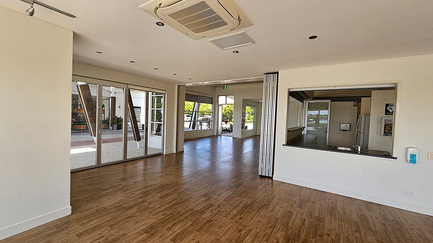 A spacious room with wooden flooring and a white ceiling featuring recessed lights and an air conditioning unit. On the left, there are large glass doors leading outside. In the centre, a hallway with more windows and an open door reveals greenery outside. On the right, a large serving window opens into a kitchen area, with a sink visible. A hand sanitiser dispenser is mounted on the wall near the window.