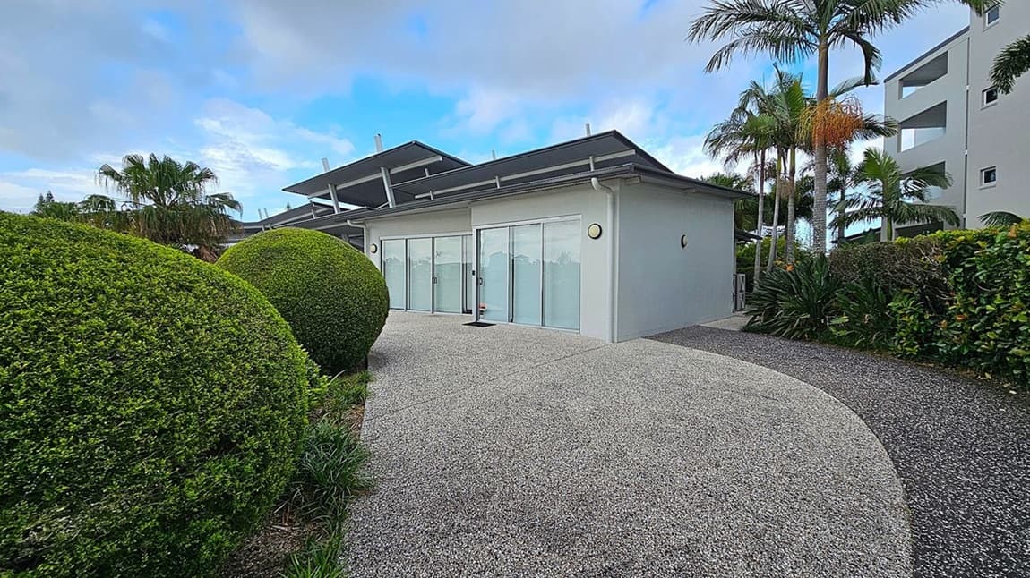 A modern building with large glass sliding doors is in the centre, surrounded by a neatly landscaped garden. On the left, there are large, round, manicured bushes. On the right, tall palm trees and dense greenery line the path. The ground is covered with a speckled grey concrete path that curves around the building. The building has a flat roof with angular extensions.