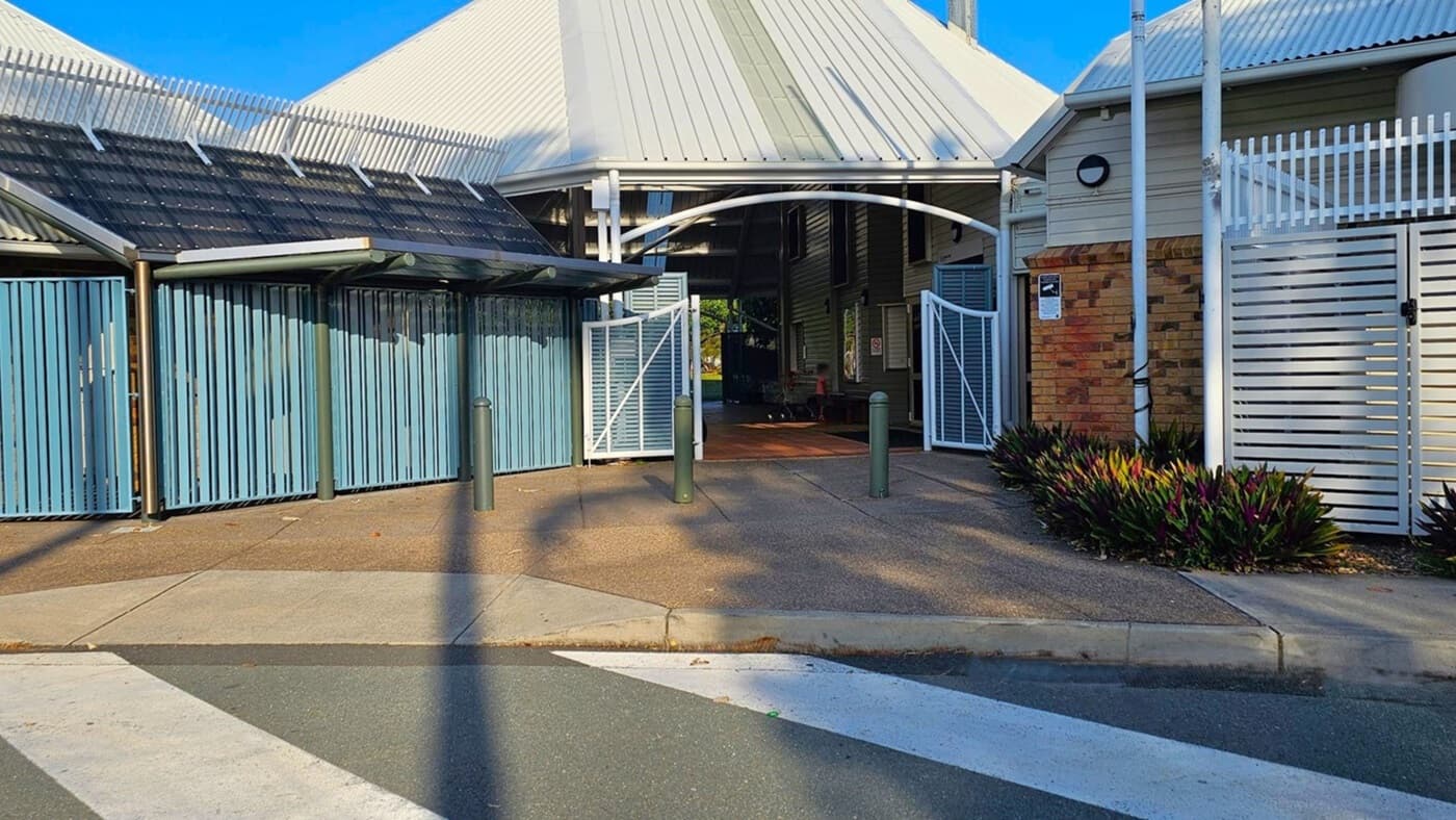 A building entrance with a white arched gate in the centre, flanked by blue vertical slatted panels on the left and a brick wall with white horizontal slats on the right. The roof is corrugated metal. There are three bollards in front of the entrance. The ground is paved, and there are plants on the right side. A pedestrian crossing is visible in the foreground.