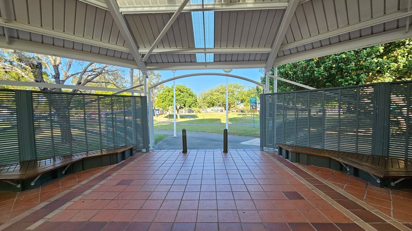 A covered outdoor area with a tiled floor and a metal roof. On the left and right, there are green slatted partitions with wooden benches attached. In the background, there is a grassy park with trees and a pathway. Two bollards are positioned at the entrance to the park.