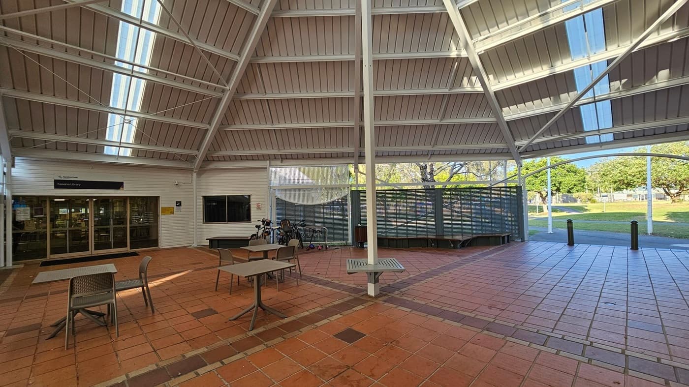 A covered outdoor area with a high, slanted roof featuring skylights. On the left, there is an entrance to a library with glass doors. In front, several tables and chairs are placed on a tiled floor. Bicycles are parked near a metal fence in the background. The right side opens to a grassy area with trees visible through the open space. The ceiling is supported by white beams.