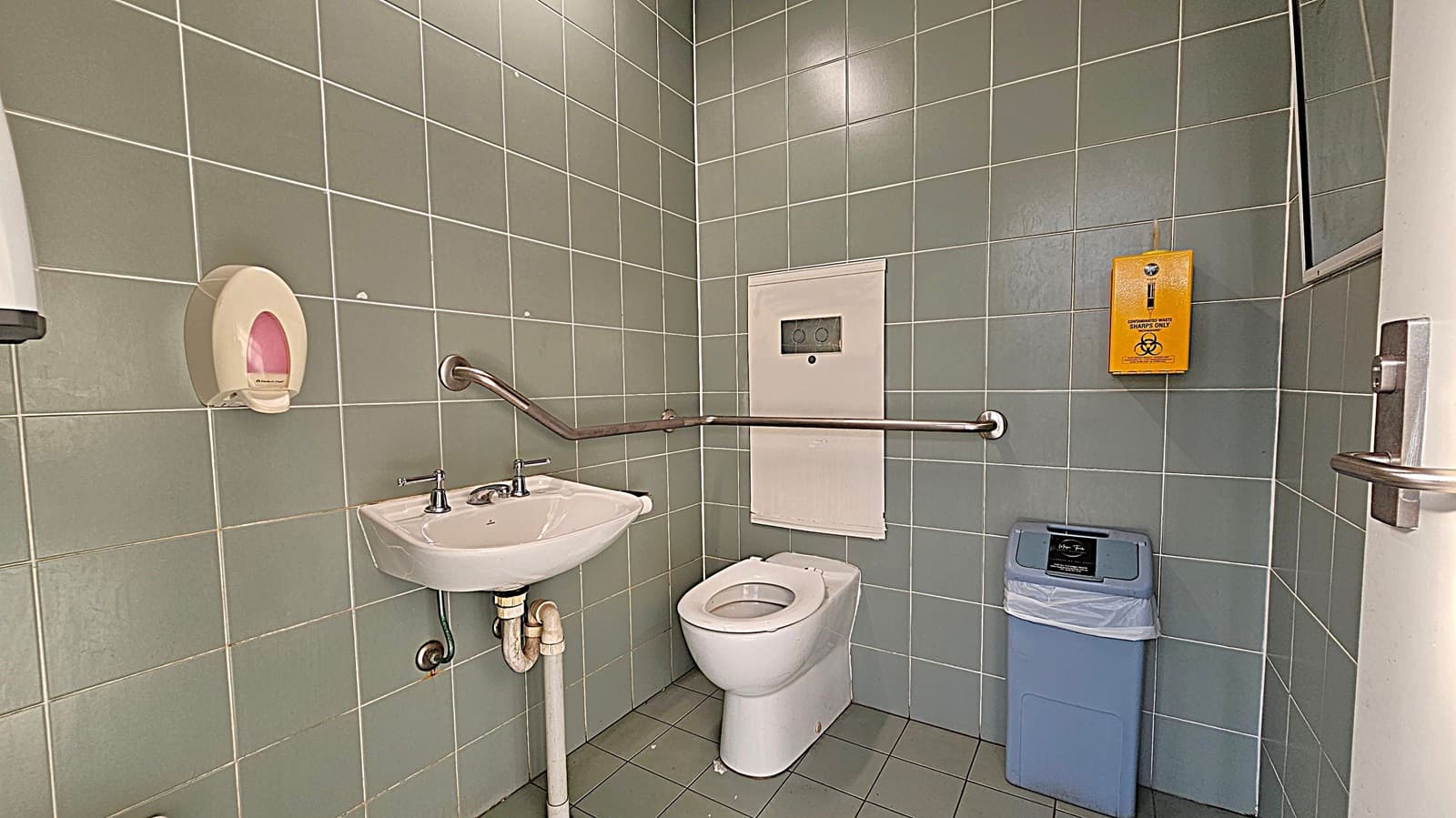 A bathroom with green tiled walls and floor. On the left, there's a white sink with a metal tap and a soap dispenser above it. A metal grab bar runs along the wall. In the centre, a white toilet is positioned beneath a panel. On the right, a yellow sharps disposal box is mounted on the wall above a blue bin. A door with a metal handle is partially visible on the far right.