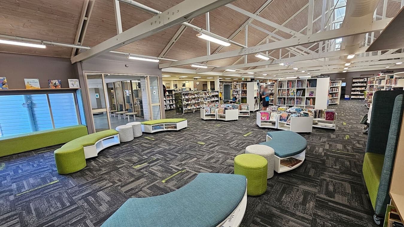 A spacious library interior with a high, wooden ceiling and exposed beams. The floor is covered with patterned carpet tiles in grey and green. On the left, there are green and grey curved benches near a large window. In the centre, there are white bookshelves filled with books, and a person is standing nearby. The right side has more bookshelves and seating areas. The background features a glass-walled room and additional bookshelves.