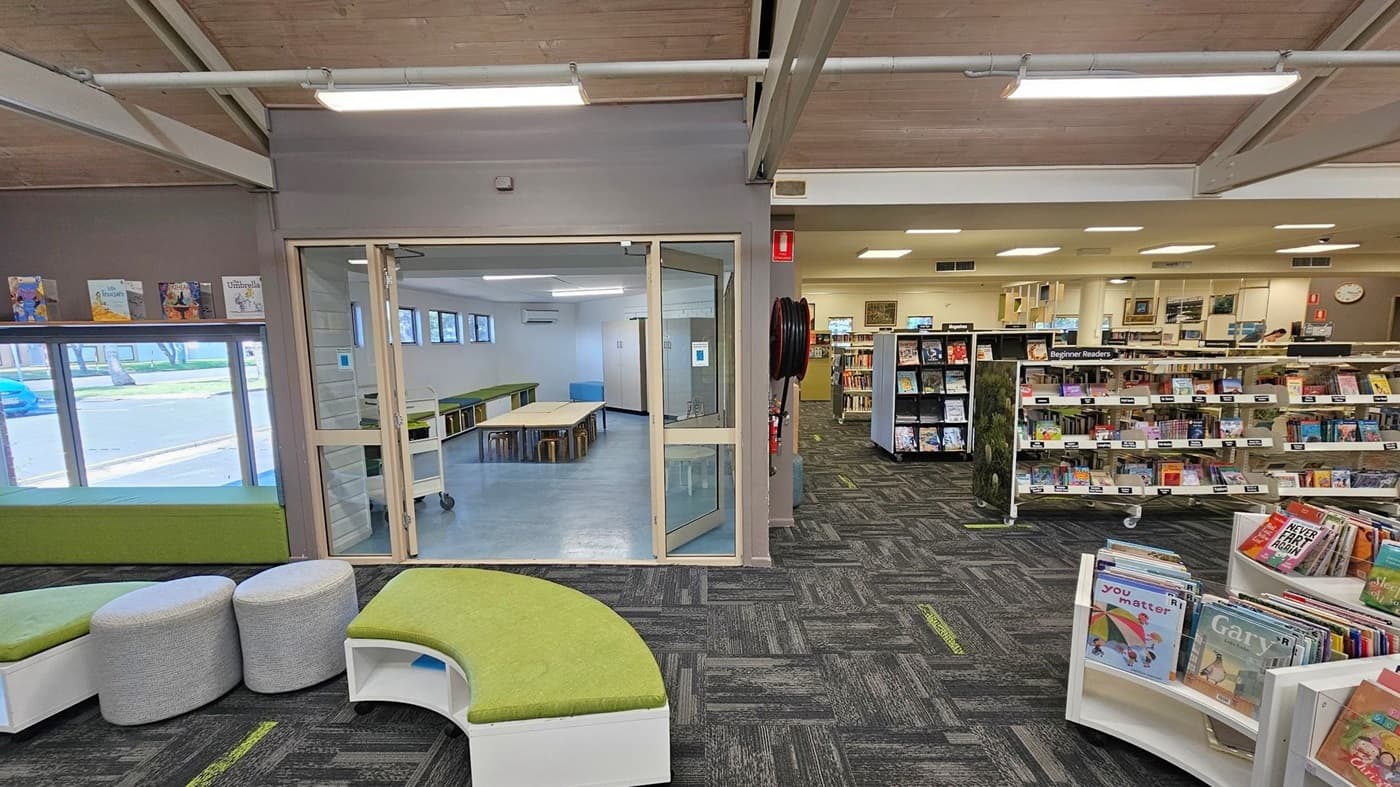 A library interior with a grey and green carpeted floor. On the left, there are green cushioned benches and round grey stools. A glass door in the centre leads to a room with tables and stools, featuring a light blue floor. On the right, there are bookshelves labelled "Beginner Readers" filled with books. The ceiling is wooden with exposed beams and fluorescent lights. A window on the left shows a view of the outside.