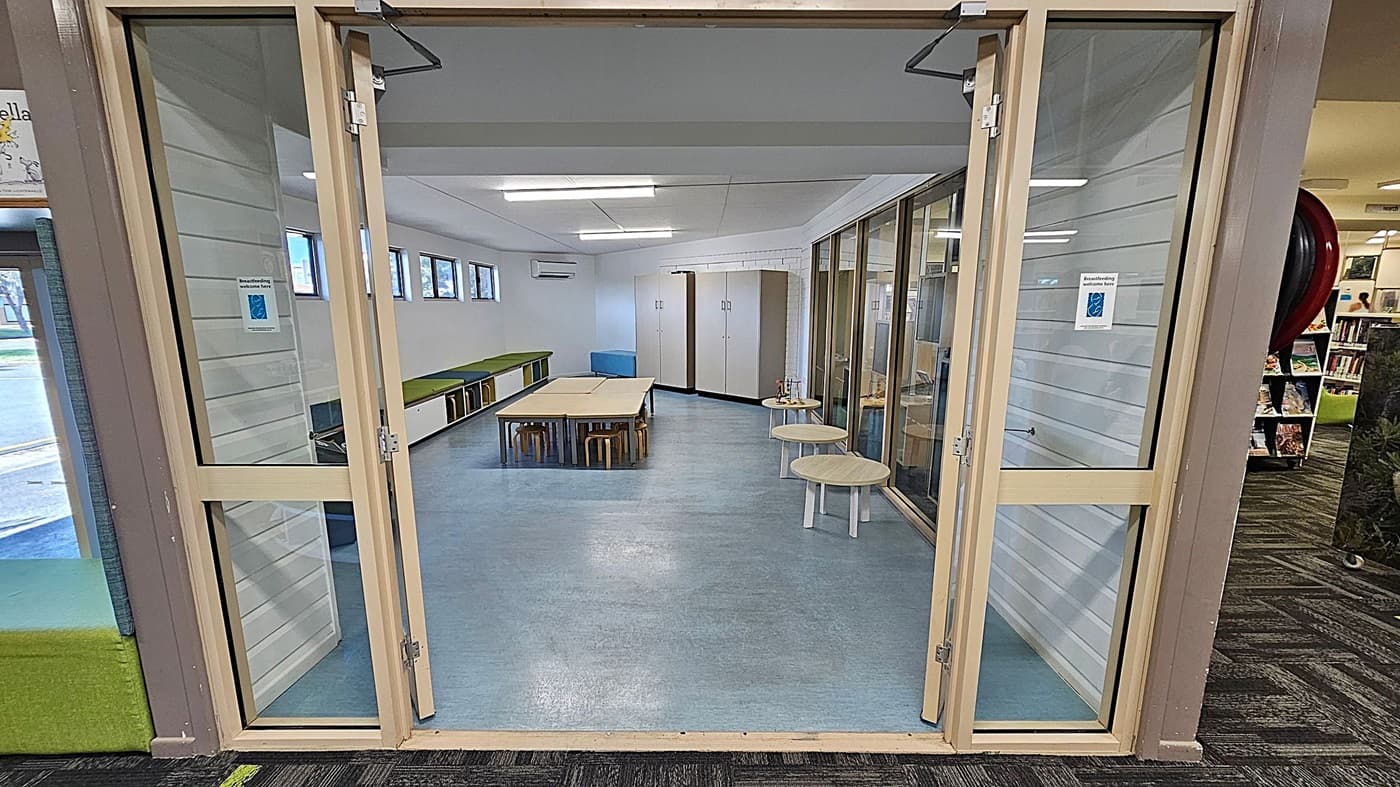 A room with glass doors and a blue floor. Inside, there are tables and stools in the centre, with green cushioned benches along the left wall. On the right, there are small round tables and a glass partition. The back wall has storage cabinets and small windows near the ceiling. The ceiling is white with fluorescent lights. The area outside the room has a patterned carpet with green and black designs.