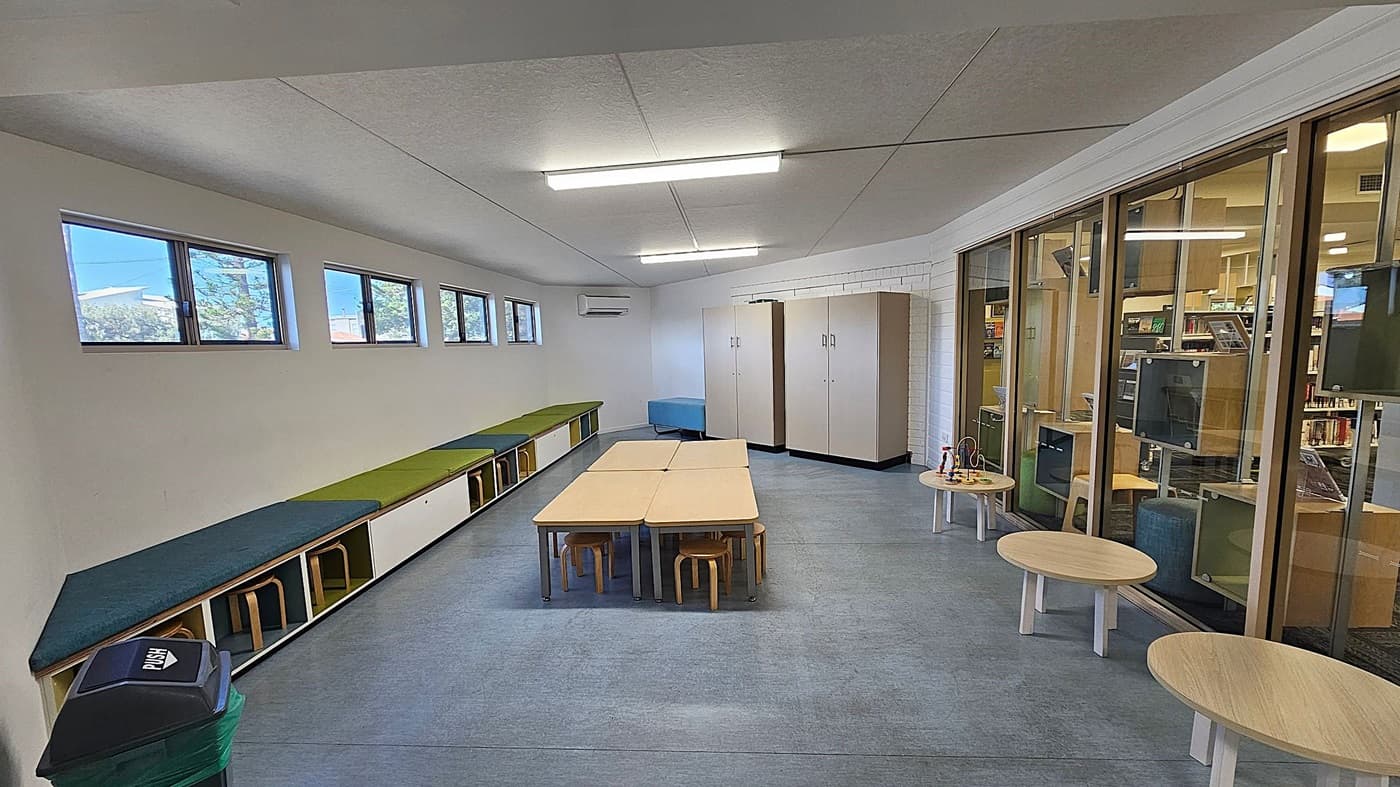 A room with a row of small windows on the left wall, above a long bench with green and blue cushions. Below the bench are stools. In the centre, there are light-coloured tables with small wooden stools. The right side features large glass panels showing another room with bookshelves. Near the glass panels are round wooden tables with toys. The floor is grey, and the ceiling has fluorescent lights. A bin is in the foreground on the left.