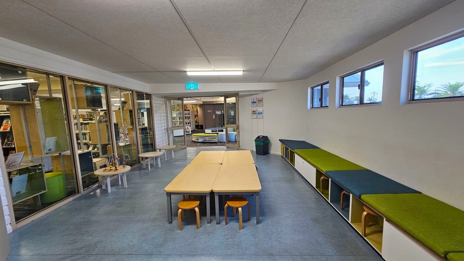 A room with a grey ceiling and blue-grey floor. On the left, there are glass walls with a view into a library, featuring bookshelves and small round tables with toys. In the centre, two rectangular tables are pushed together with small wooden stools underneath. On the right, a long bench with green and blue cushions sits below a row of small windows. A green and black bin is near the entrance, which leads to another room with more bookshelves.