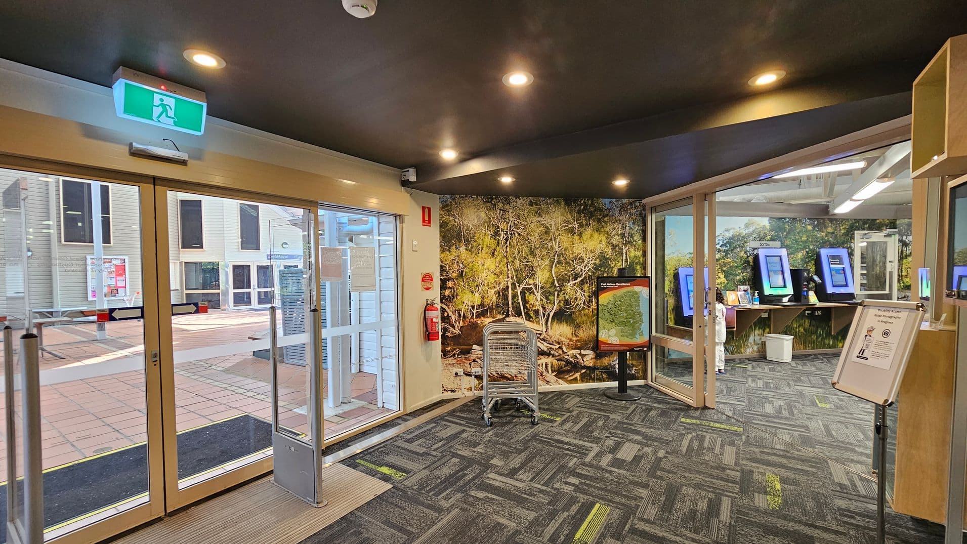 Glass entrance doors to a library with a "Zero Tolerance" sign on the left and opening hours on the right. Inside, bookshelves are visible with books and displays. The floor is tiled outside and carpeted inside. The ceiling has recessed lighting.