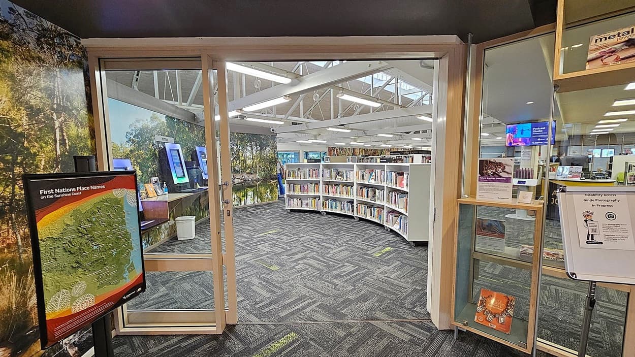 A library entrance with glass doors. On the left, a sign displays "First Nations Place Names on the Sunshine Coast" with a map. Behind it, a wall features a nature mural. Inside, curved bookshelves are filled with books. On the right, a glass display case holds informational materials, and a sign mentions "Disability Access Guide Photography in Progress." The floor is carpeted with a grey and green pattern, and the ceiling has exposed white beams with fluorescent lights.