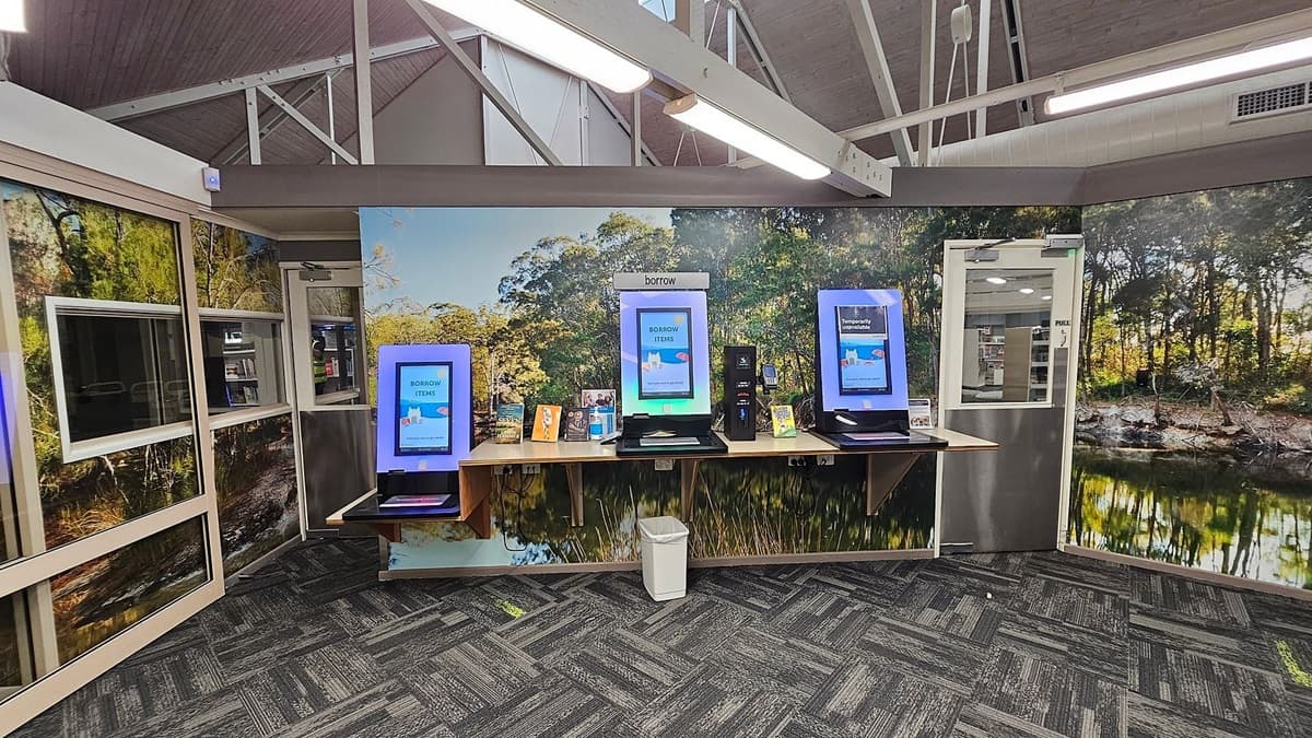 A library interior with three self-service kiosks on a wooden counter against a wall featuring a nature mural. The kiosks display screens with "BORROW ITEMS" and "Temporarily Unavailable" messages. Below the counter is a white bin. The floor is carpeted with a grey and green pattern. The ceiling has exposed beams and fluorescent lights. On the left, there are windows with a view of the library interior, and on the right, a door with a "PULL" sign.