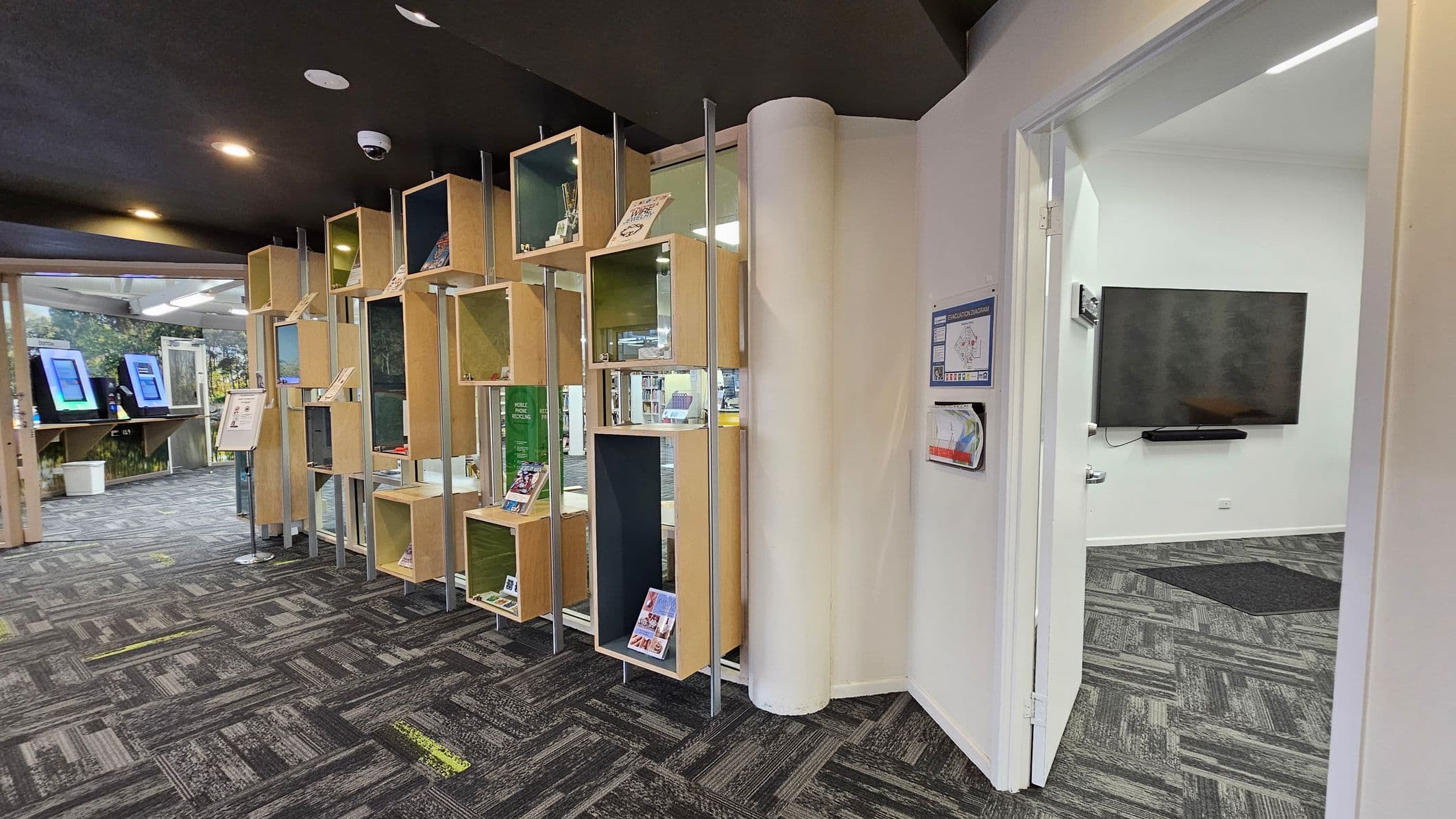 On the left, there are wooden display shelves with various items and books. Behind them, a section with digital screens is visible. The floor is carpeted with a grey and green pattern. On the right, an open door leads to a room with a large wall-mounted TV. The ceiling is dark with recessed lighting.