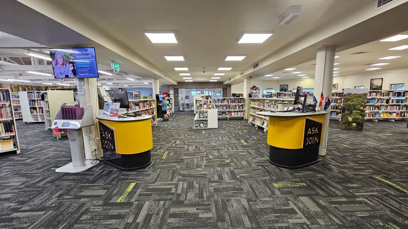 A library interior with a high, wooden ceiling supported by white beams. On the left, there are curved white bookshelves filled with books, labelled "Food and Drink." The floor is covered with dark patterned carpet tiles. On the right, there's a doorway with wooden shelving units. In the background, more bookshelves and a yellow circular desk are visible, along with a digital screen displaying content. The space is well-lit with fluorescent lights.