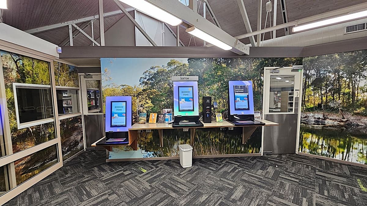 A library interior with three self-service kiosks on a wooden counter against a wall featuring a nature mural. The kiosks display screens with "BORROW ITEMS" and "Temporarily Unavailable" messages. Below the counter is a white bin. The floor is carpeted with a grey and green pattern. The ceiling has exposed beams and fluorescent lights. To the right, a glass door leads to another room with visible bookshelves.