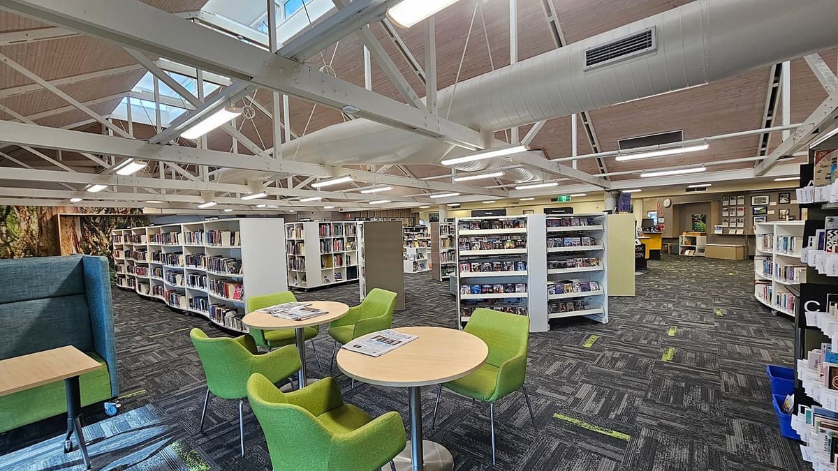 A library interior with a high ceiling featuring exposed white beams and ductwork. In the foreground, there are round tables with green chairs. The floor is carpeted in a dark pattern with green accents. Bookshelves filled with books and media are in the middle and background. On the left, there's a green cushioned seating area. The back wall has framed pictures and a clock. Skylights provide natural light from above.