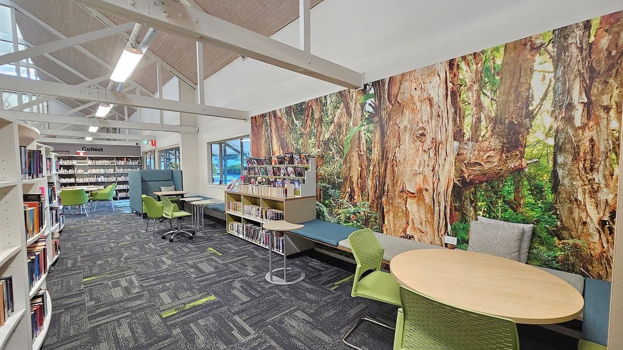 Library interior with a high, sloped ceiling featuring exposed beams. On the right, a large mural of a forest scene covers the wall, with a cushioned bench and a small round table with green chairs in front. In the middle, bookshelves display various books and magazines. On the left, more bookshelves and a seating area with green chairs and tables. The floor is carpeted with a grey and green pattern, and large windows let in natural light.