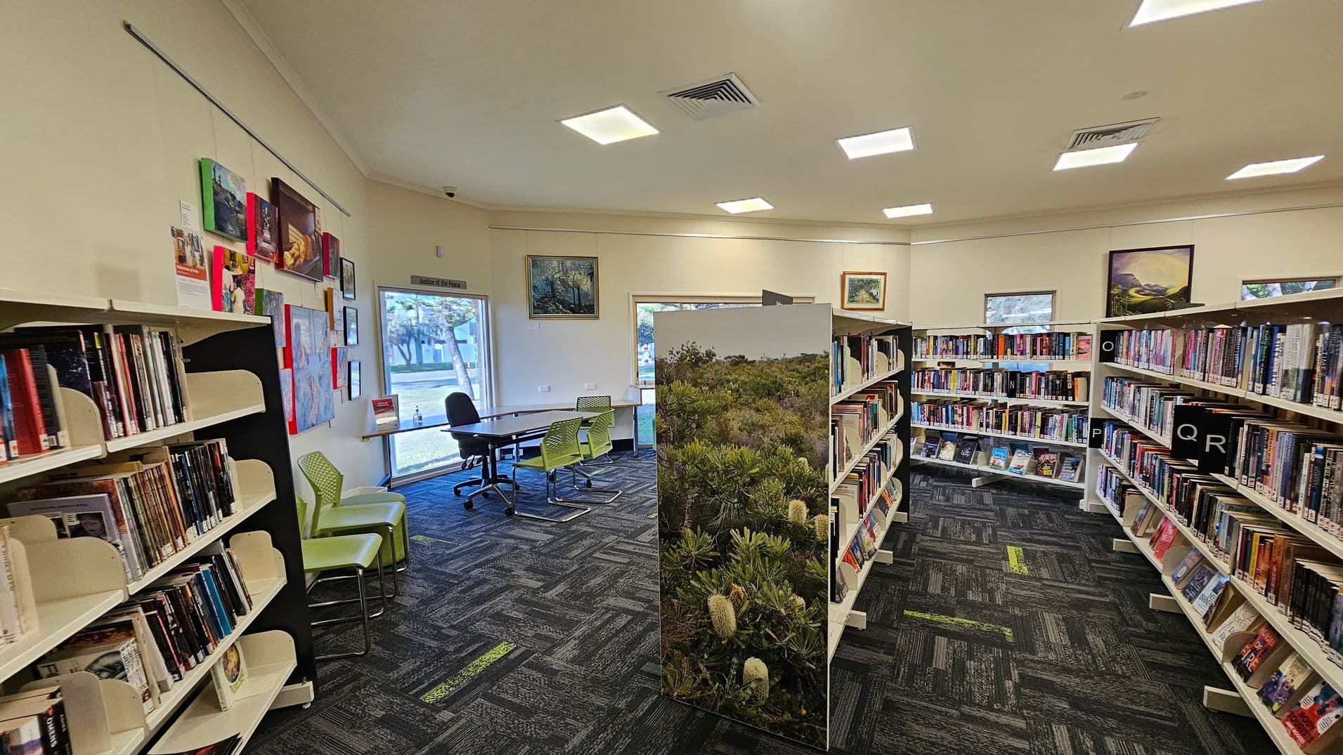 A library interior with bookshelves on the left and right, filled with books. In the middle, a partition with a nature scene separates the space. The floor is carpeted with a dark pattern featuring green accents. On the left wall, there are colourful artworks and a window with a view outside. In the background, a table with chairs is near another window. The ceiling has recessed lighting.