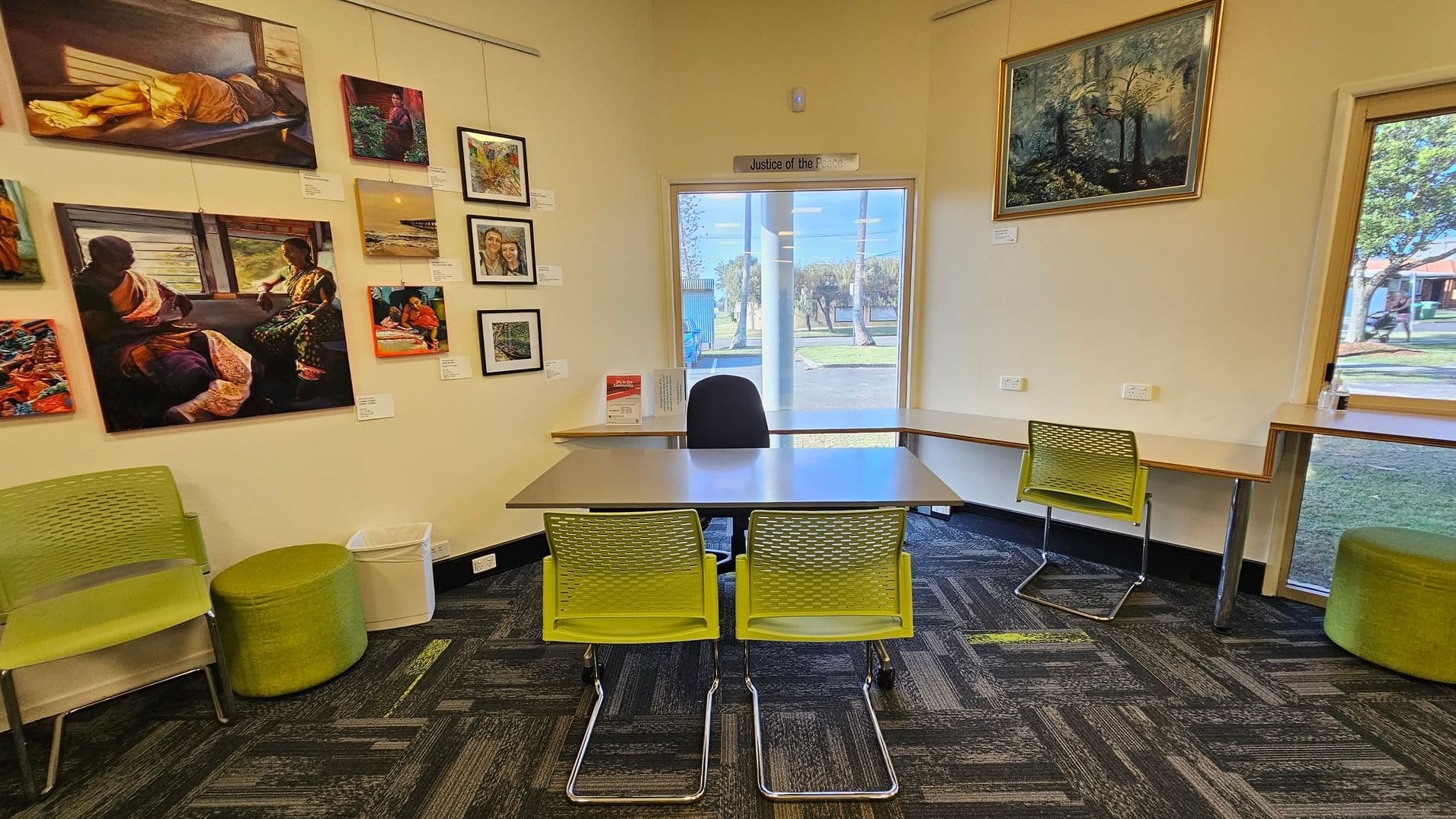 A library interior with bookshelves on both sides filled with books. In the middle, a partition displays an image of greenery. The left side has a wall with artwork and a window, next to which are tables and green chairs. The floor is carpeted with a dark pattern featuring green accents. The ceiling has recessed lighting.