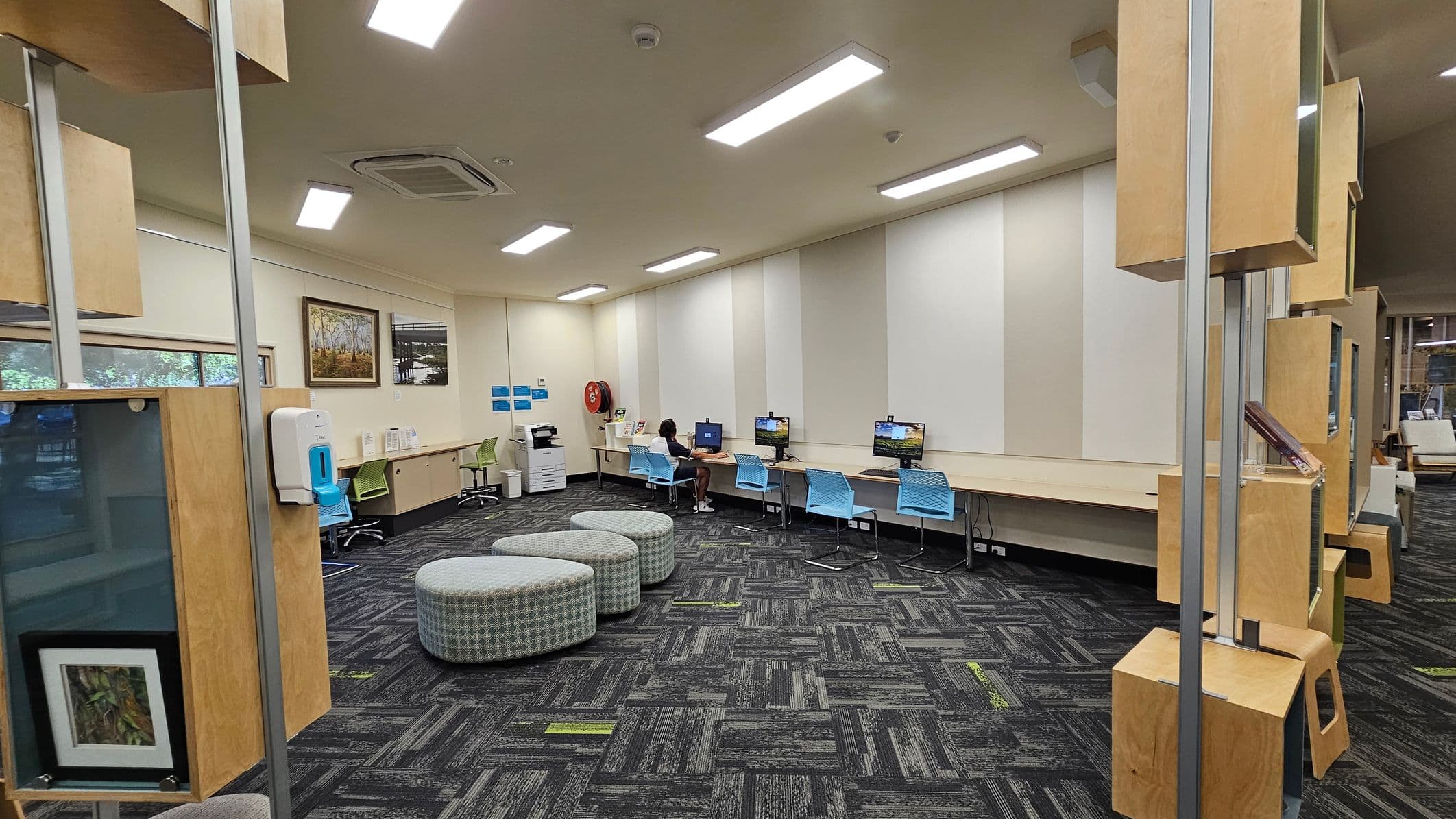A room with a patterned carpet floor and cream walls. On the left, there are several colourful paintings and photographs displayed. Below them, a green chair and a round green ottoman are next to a white bin. In the centre, a desk with two green chairs faces a window labelled "Justice of the Peace." Behind the desk is another chair. On the right, a framed painting hangs above a long counter with a green chair. Large windows provide a view of trees and grass outside.
