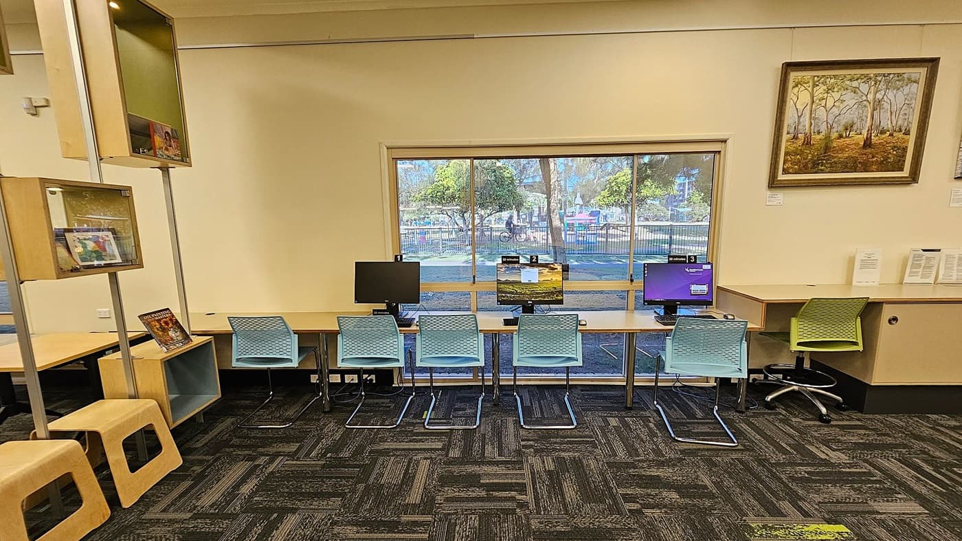 A room with a row of three computers on a long desk against a large window. Each computer has a blue chair in front of it. To the right, there's a green chair at a separate desk with papers on it. On the left, there are wooden shelves with books and a couple of wooden stools. A framed painting of trees hangs on the wall to the right. The floor is covered with a patterned carpet in shades of green and grey.