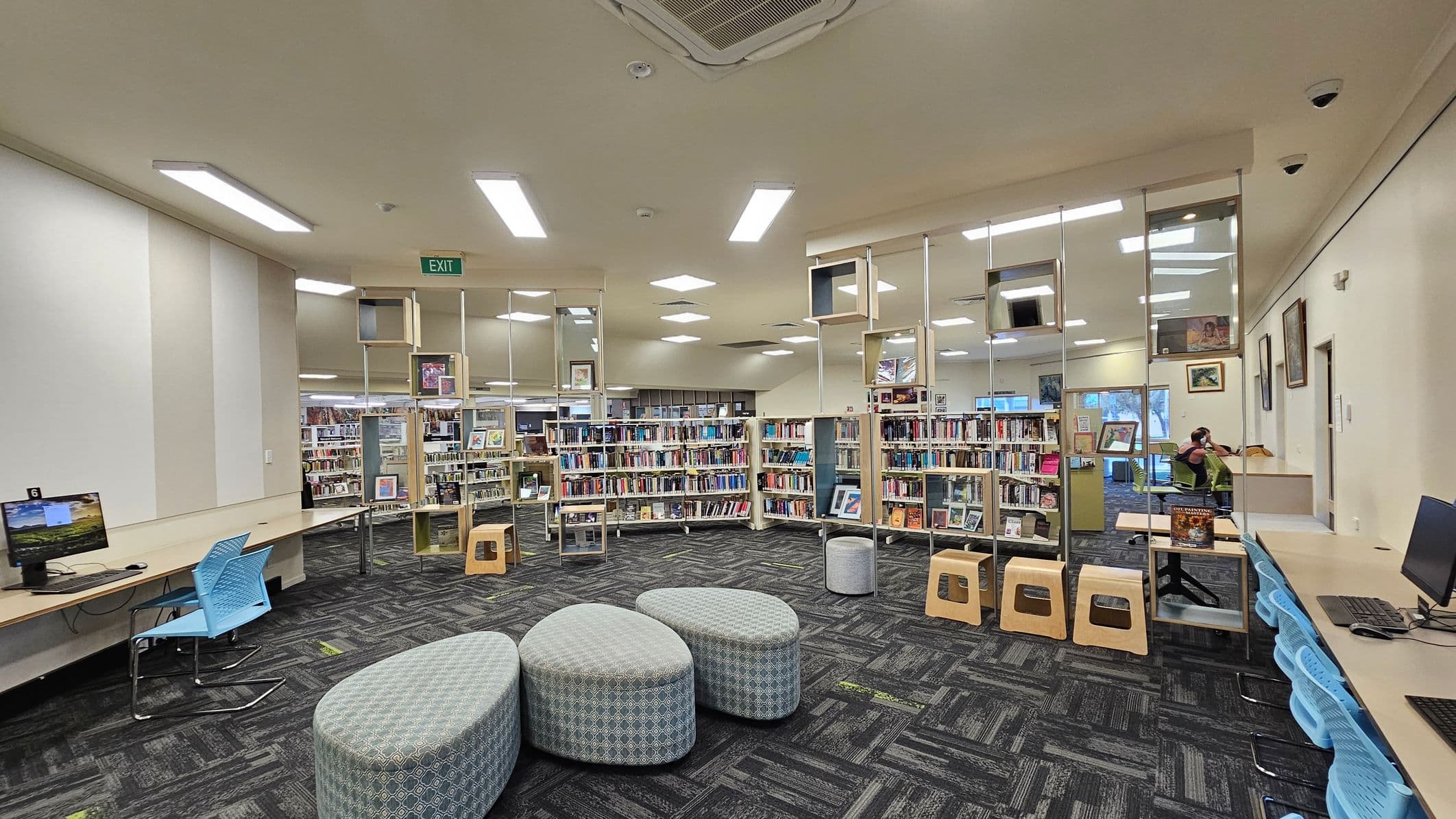 A library interior with a patterned carpet floor and a ceiling with recessed lighting. In the foreground, there are three grey, oval-shaped cushioned seats. On the left, a long desk with computers and blue chairs. In the centre, bookshelves filled with books and decorative cube shelves above. On the right, more desks with computers and a person sitting at one. The walls have framed pictures, and an exit sign is visible at the back.