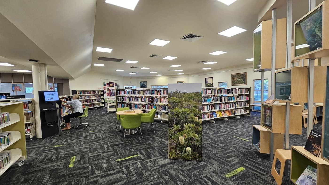 A library interior with a patterned carpet and a ceiling with square lights. On the left, a person is seated at a computer station. In the centre, there are bookshelves filled with books and a round table with green chairs. A large panel with a nature image stands nearby. On the right, there are wooden display shelves with books and framed pictures. Windows are visible in the background, allowing natural light in.