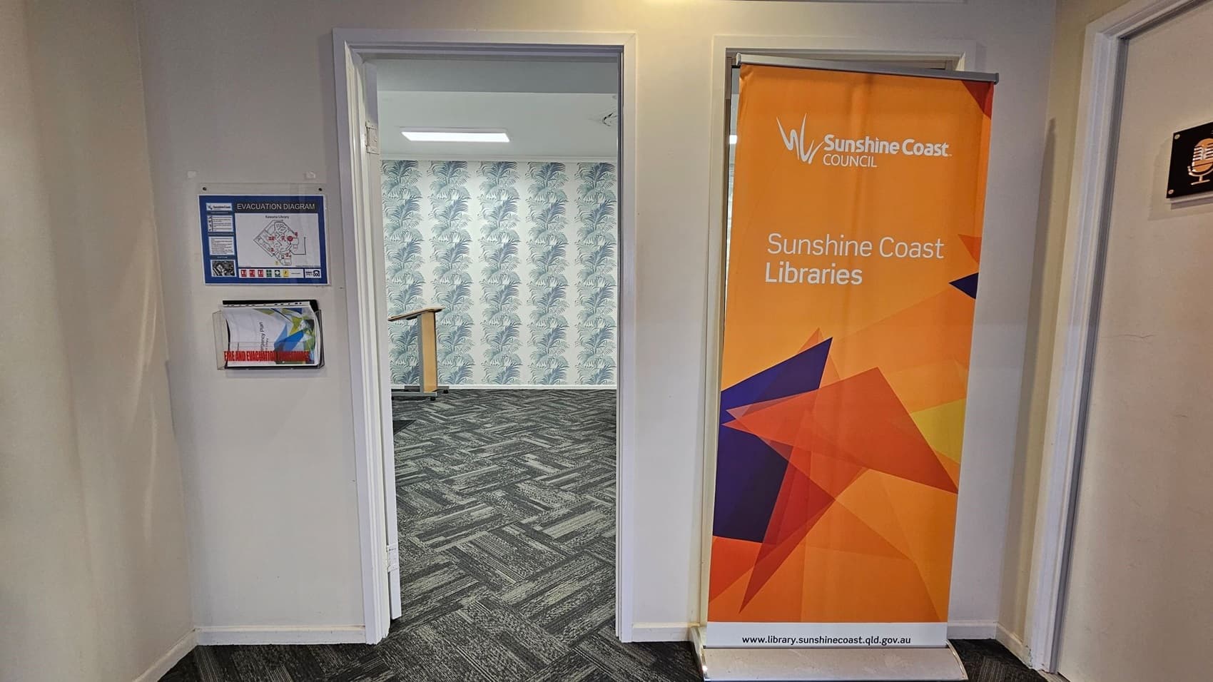 On the left, there are wooden display shelves with various items and informational signs. In the background, there are digital kiosks against a wall with a nature-themed mural. On the right, an open door leads to a room with a large flat-screen TV mounted on a white wall. The floor is covered with a patterned carpet in shades of grey and green. The ceiling is dark with recessed lighting.