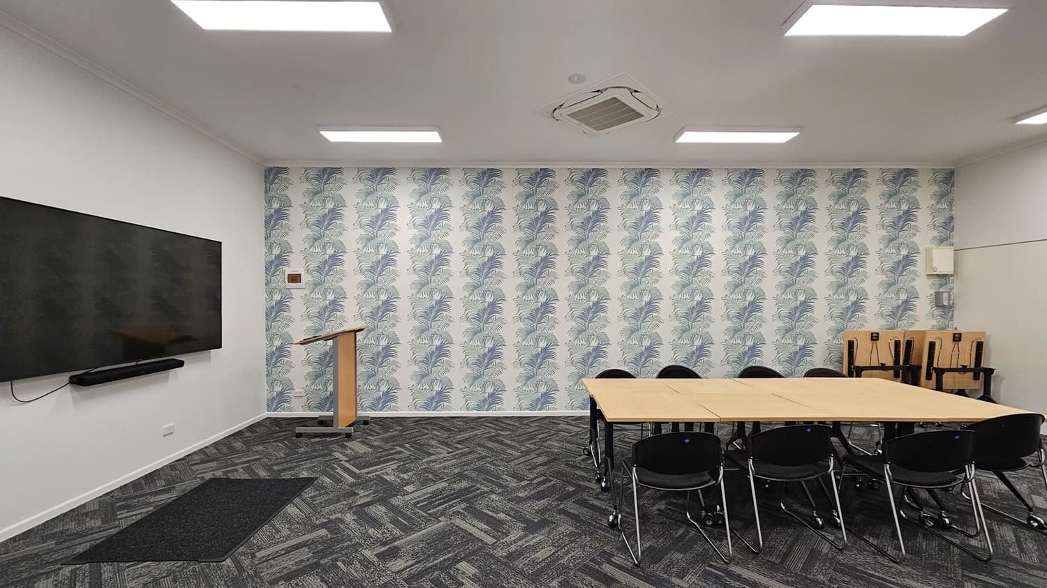 A room with a patterned wallpaper featuring leaf designs on the back wall. On the left, a large flat-screen TV is mounted on a white wall above a black mat. A wooden podium stands near the centre. On the right, a rectangular table with black chairs is arranged, and several stacked chairs are in the corner. The floor is covered with dark patterned carpet tiles, and the ceiling has recessed lighting and an air conditioning unit.