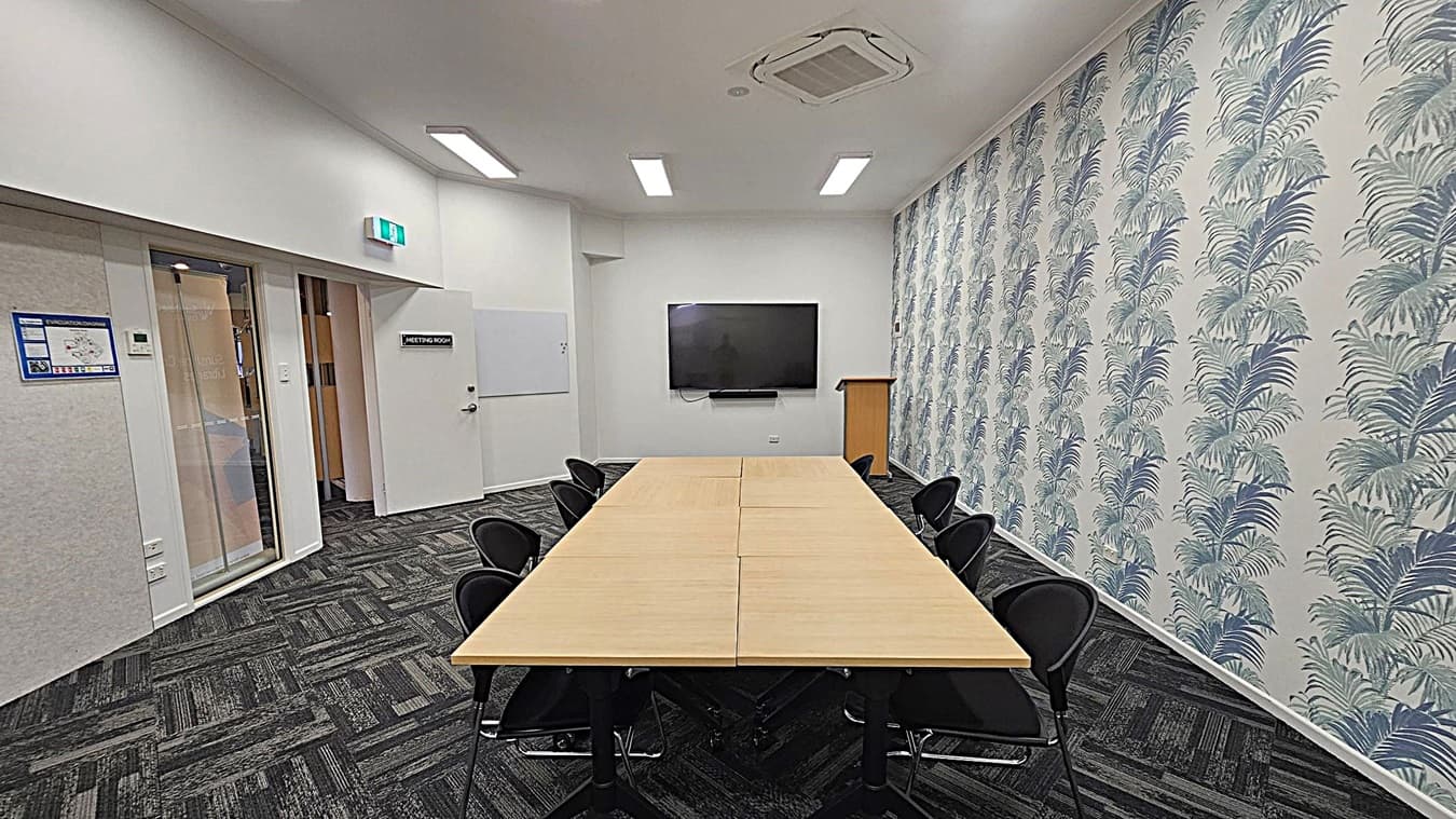 A meeting room with a rectangular wooden table in the centre, surrounded by black chairs. The right wall features a patterned wallpaper with palm leaves. At the far end, a large flat-screen TV is mounted on the wall above a wooden lectern. The left side has a door labelled "Meeting Room" and a glass panelled door. The floor is covered with dark patterned carpet tiles, and the ceiling has recessed lighting and an air conditioning unit.